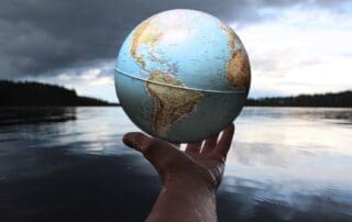 person holding a globe in the palm of their hand with a lake in the background