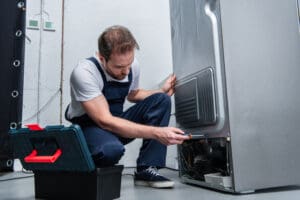 adult bearded repairman in working overall fixing refrigerator by screwdriver in kitchen adult bearded repairman in working overall fixing refrigerator by screwdriver in kitchen
